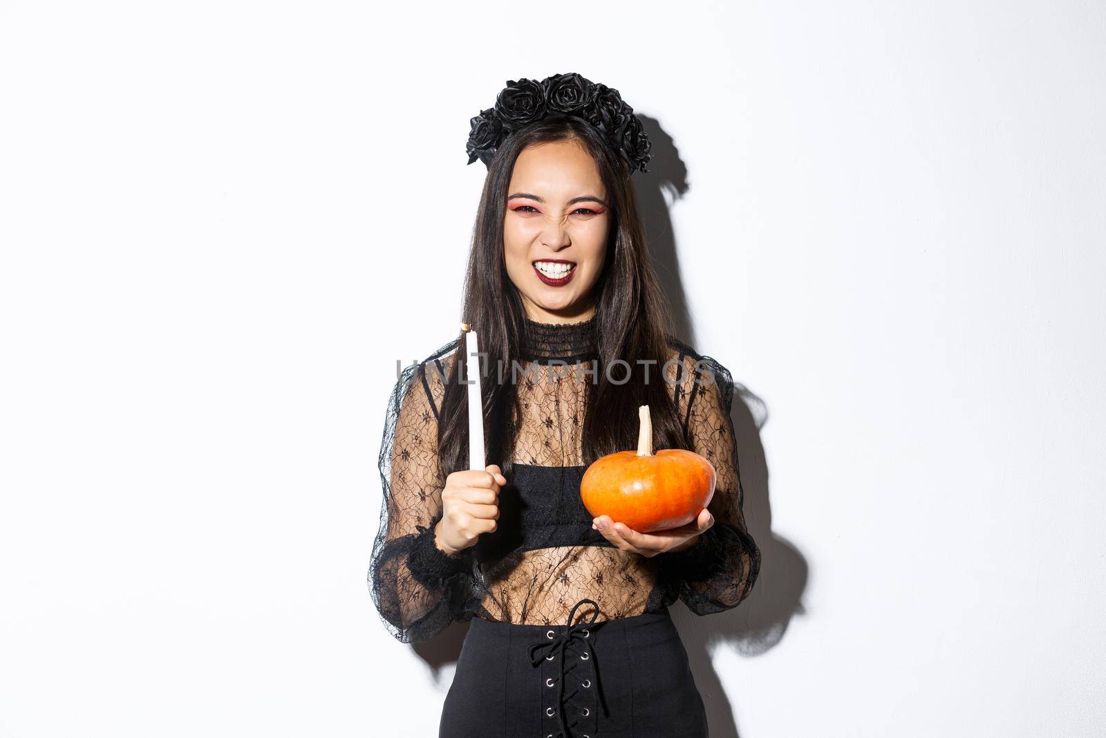 Image of asian evil witch in gothic lace dress and black wreath, laughing and grimacing, holding candle with pumpkin, celebrating halloween.