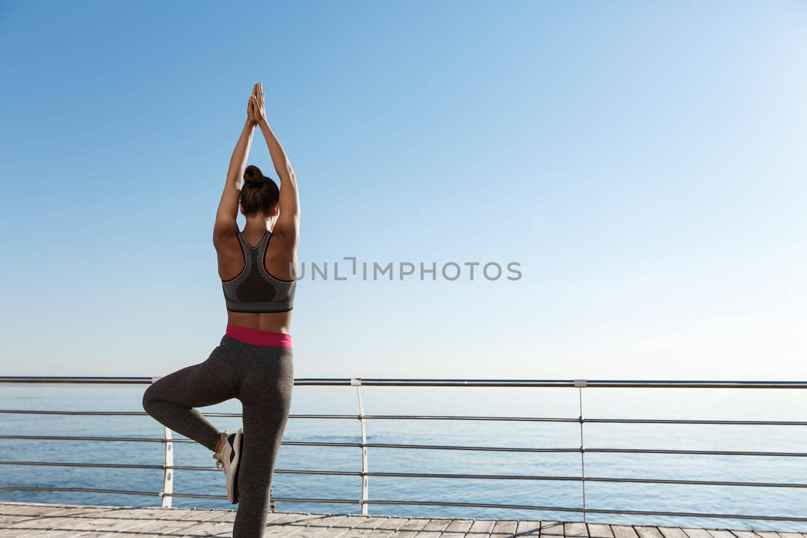 Rear view of fit and healthy woman doing yoga exercises on a pier, looking at the sea during workout.