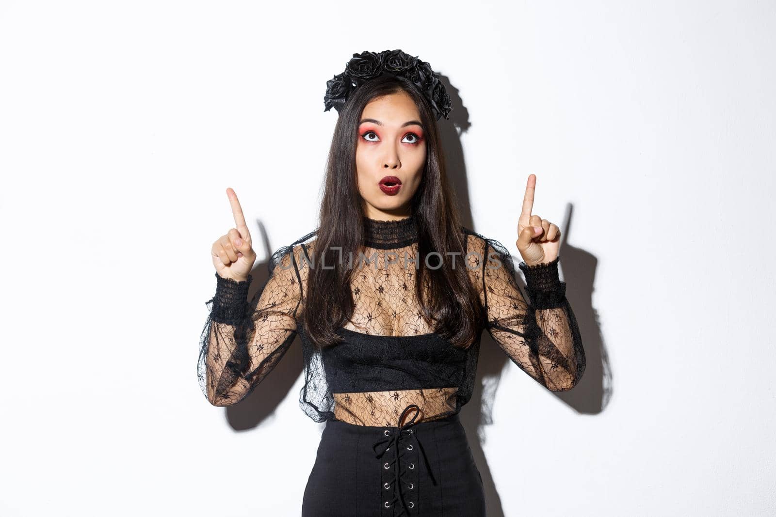 Surprised attractive asian woman in witch outfit pointing fingers up, showing halloween banner, promo of party. Beautiful female in black gothic dress looking amazed.