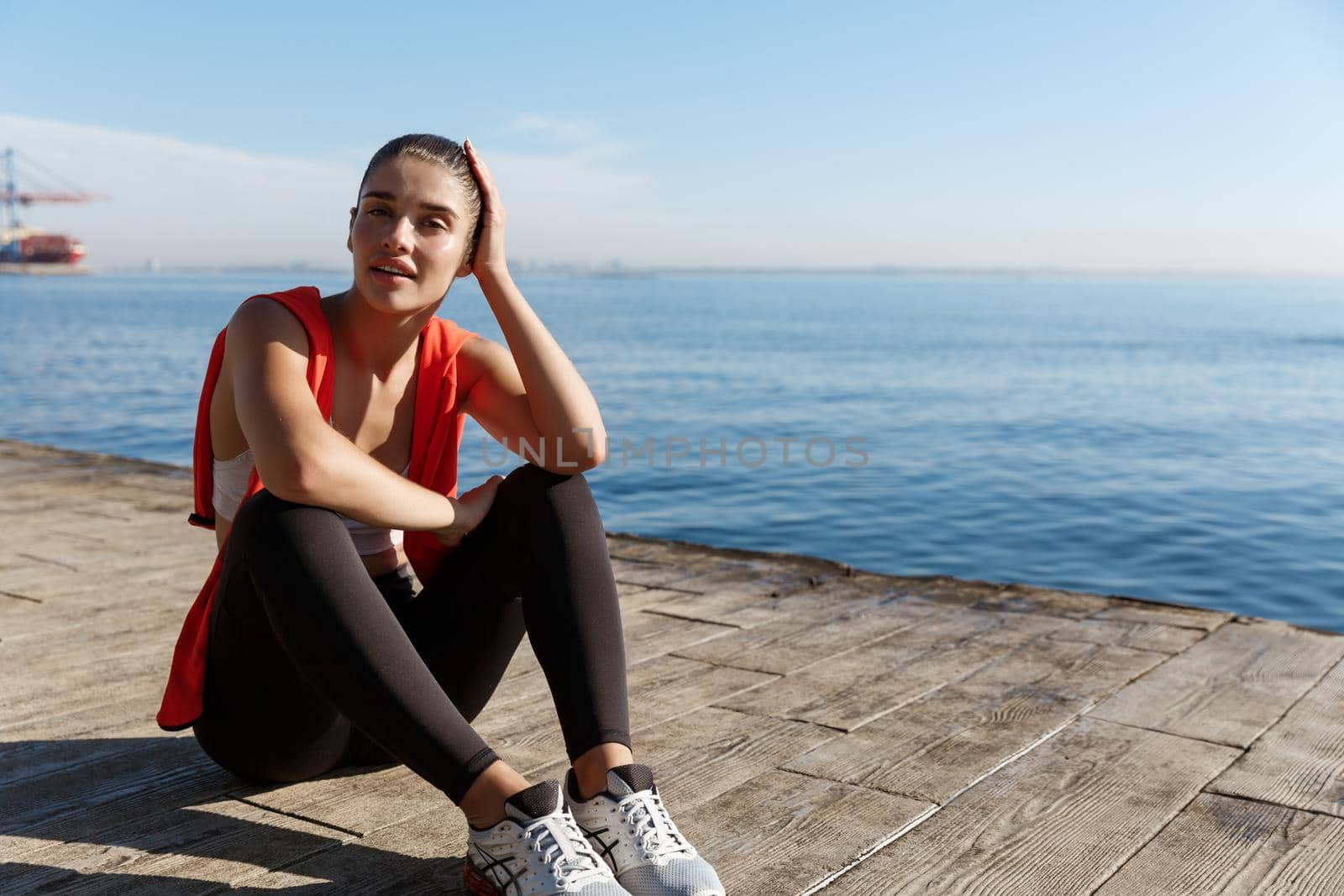 Outdoor shot of attractive fitness woman having a break after workout or jogging, sitting near sea and contemplating the view.