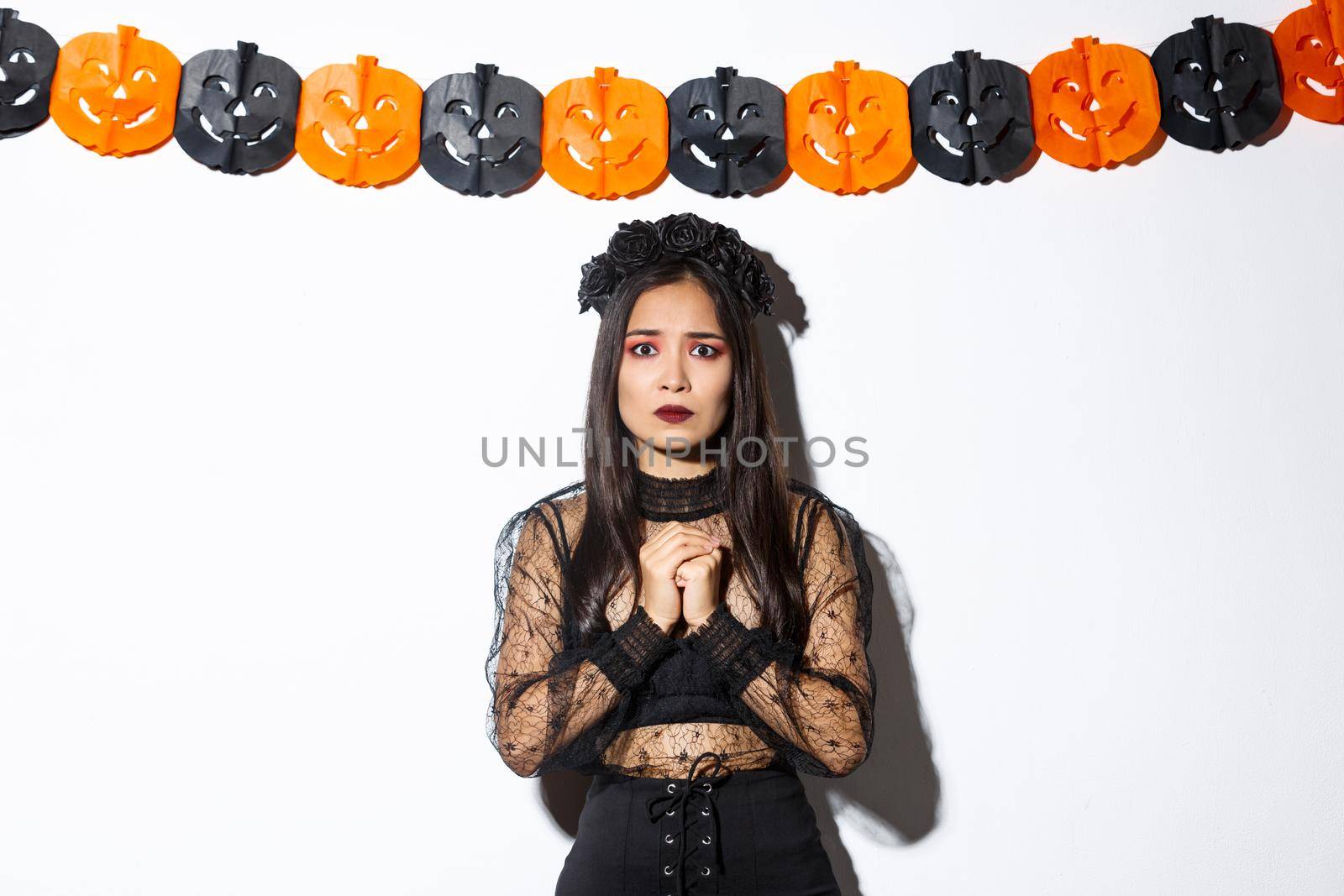 Image of scared and worried asian woman in witch costume looking concerned, wearing witch costume and standing against pumpkin banners.
