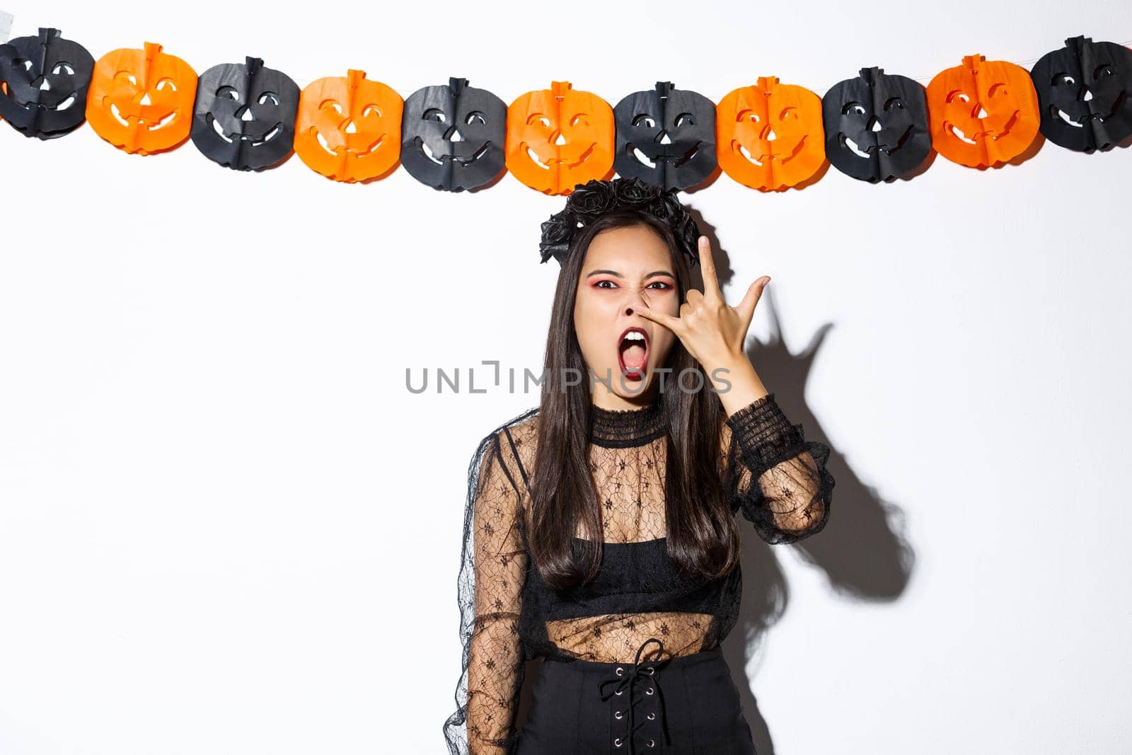Image of funny asian woman in witch costume grimacing, wearing gothic lace dress and standing against pumpkin streamers, decorations for halloween by Benzoix