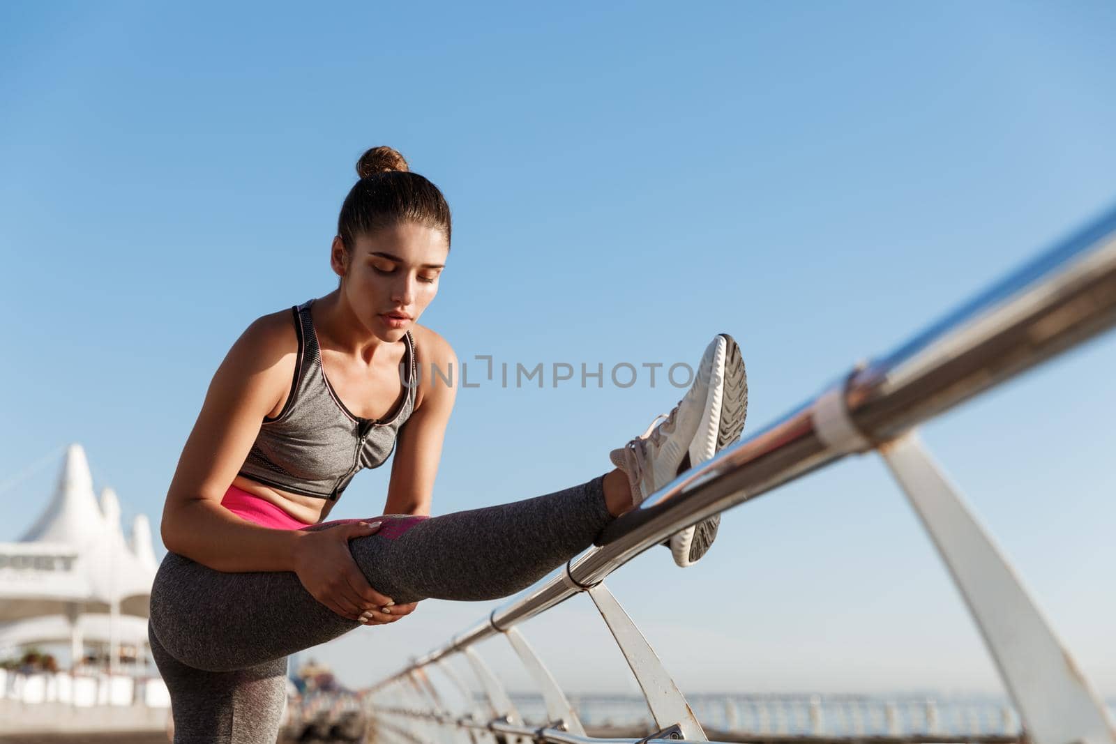 Young attractive female runner warming-up before training outdoors. Sportswoman stretching leg, prepare for jogging on the seaside promenade by Benzoix