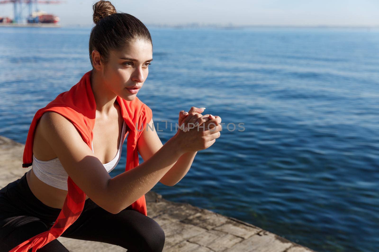 Close-up of focused young female athlete workout on a pier, doing squats and looking at sea.