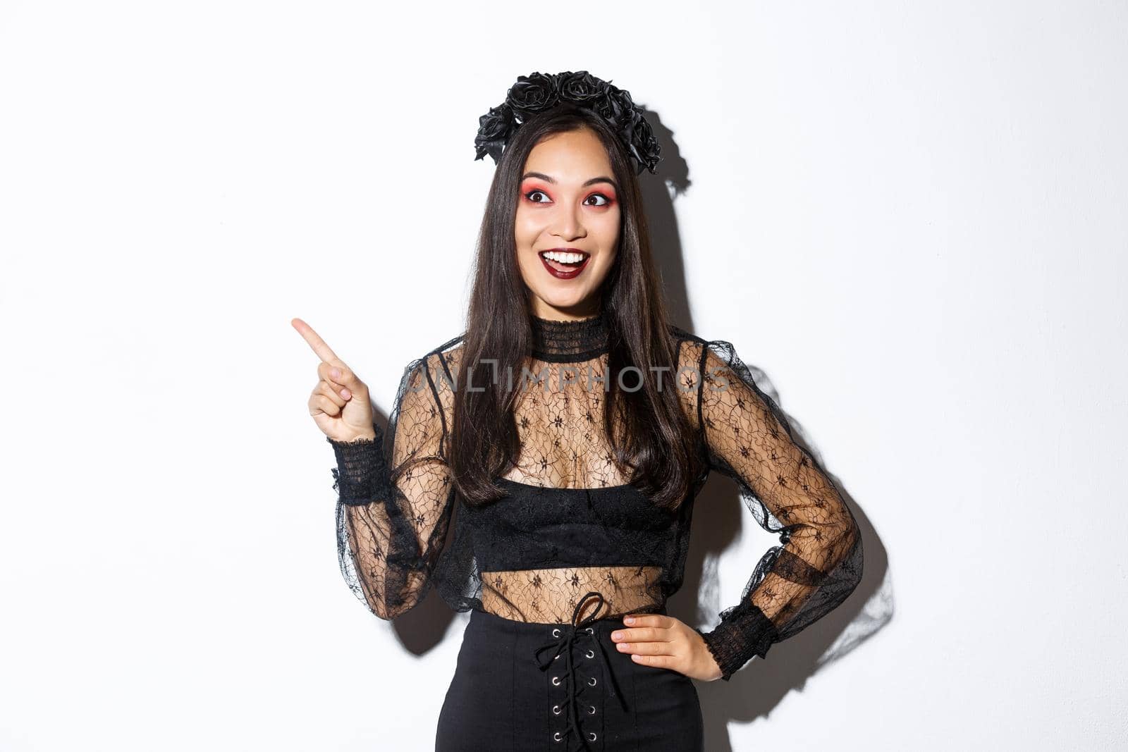 Excited happy asian woman in black lace dress and wreath looking amazed at upper left corner, pointing finger at your halloween promo banner, standing over white background.