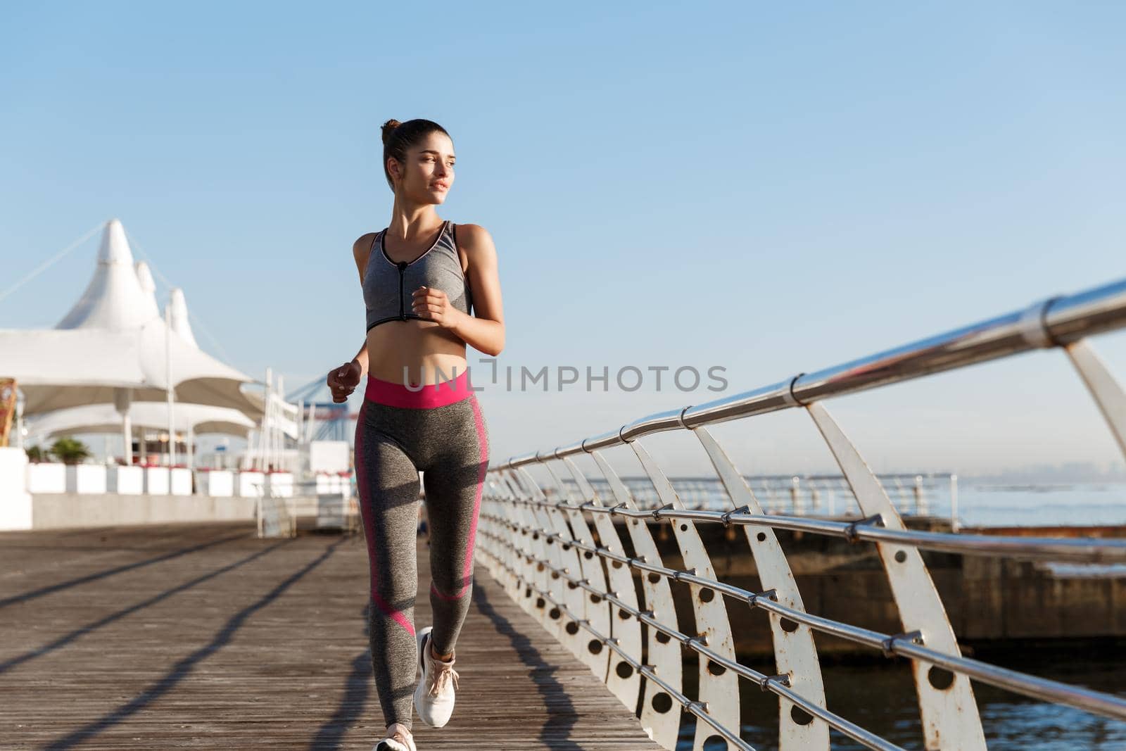 Full length shot of beatiful and healthy woman running along the seaside promenade, looking at sea during workout. Sportswoman training on pier, jogging alone.