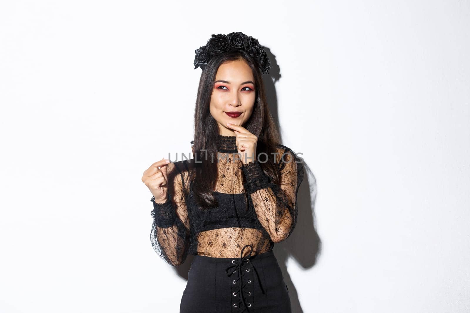 Image of smiling beautiful asian woman in gothic lace dress and wreath, thinking while holding credit card, standing over white background.