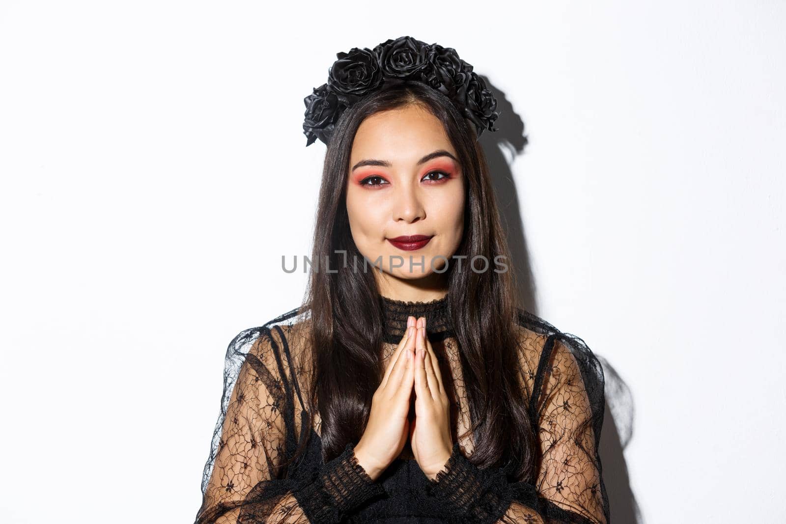 Close-up of young asian woman in black gothic dress and wreath holding hands in pray, girl wearing witch costume and celebrating halloween by Benzoix