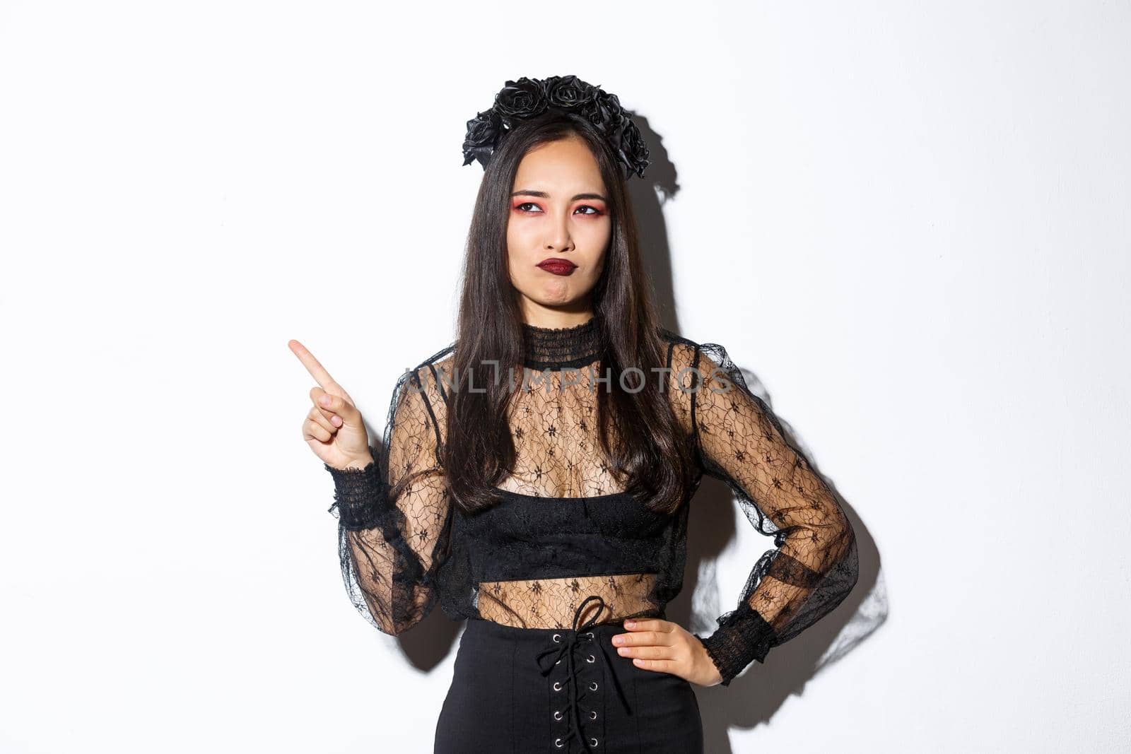 Unamused and skeptical asian beautiful woman in witch dress, looking at upper left corner with displeased smirk, standing over white background, showing logo or promo banner.