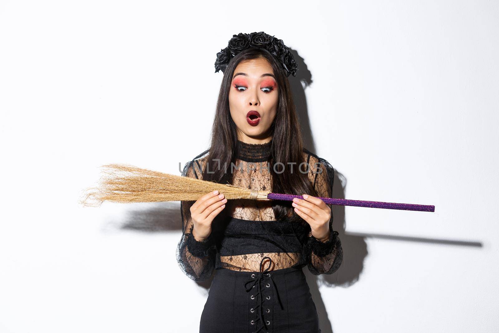 Image of amazed young witch looking at broom with surprised expression. Concept of halloween by Benzoix