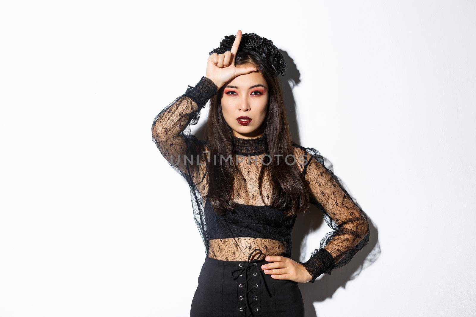 Image of arrogant sexy asian woman in witch costume showing loser gesture on forehead, mocking someone at halloween party, standing over white background.