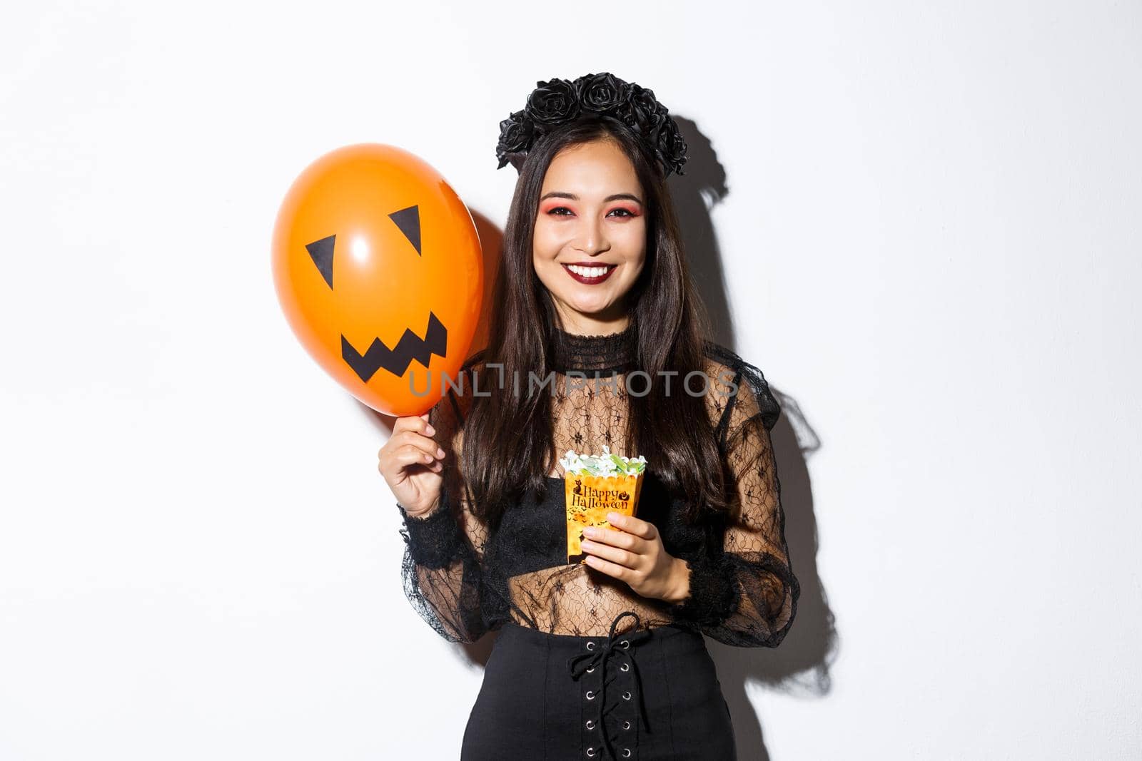 Image of cheerful asian girl trick or treating on halloween, holding sweets and orange balloon with scary face by Benzoix
