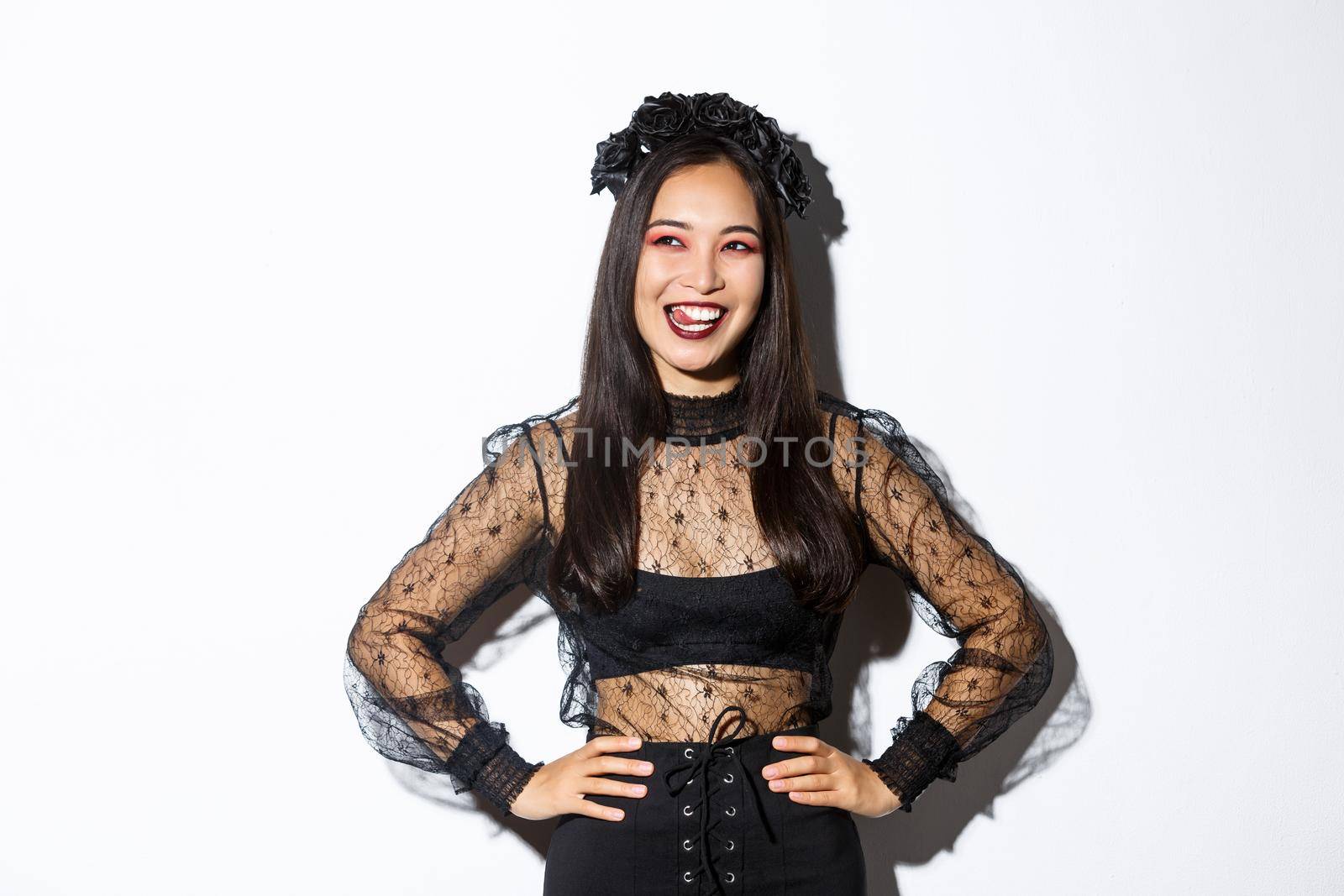 Image of carefree smiling asian woman in gothic dress and black wreath looking thoughtful at upper left corner, licking lips from desire or temptation, standing over white background by Benzoix