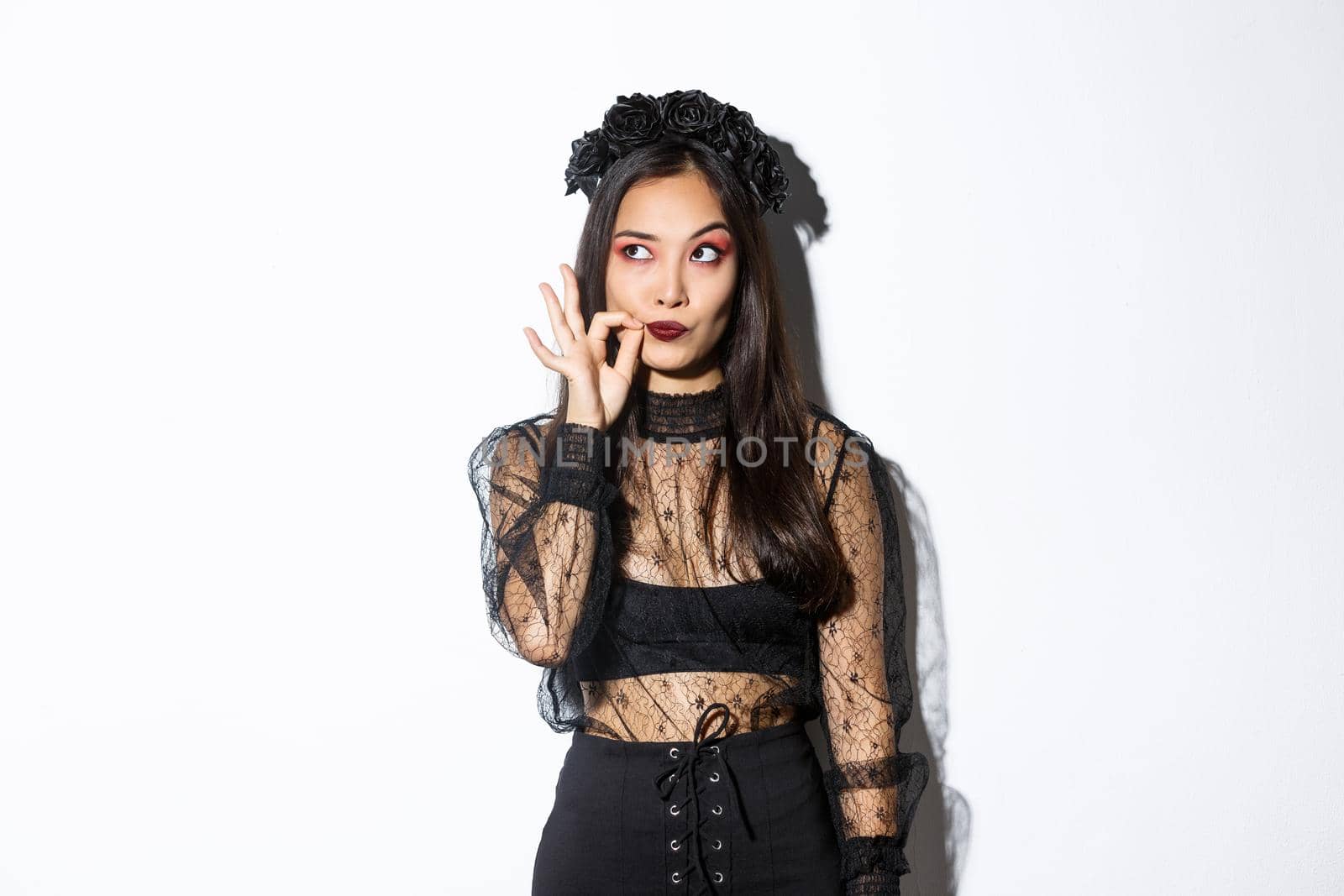 Young gothic woman in black lace dress, dressed-up like witch for halloween party, making seal on lips, zipping mouth as making promise keep secret, standing over white background.