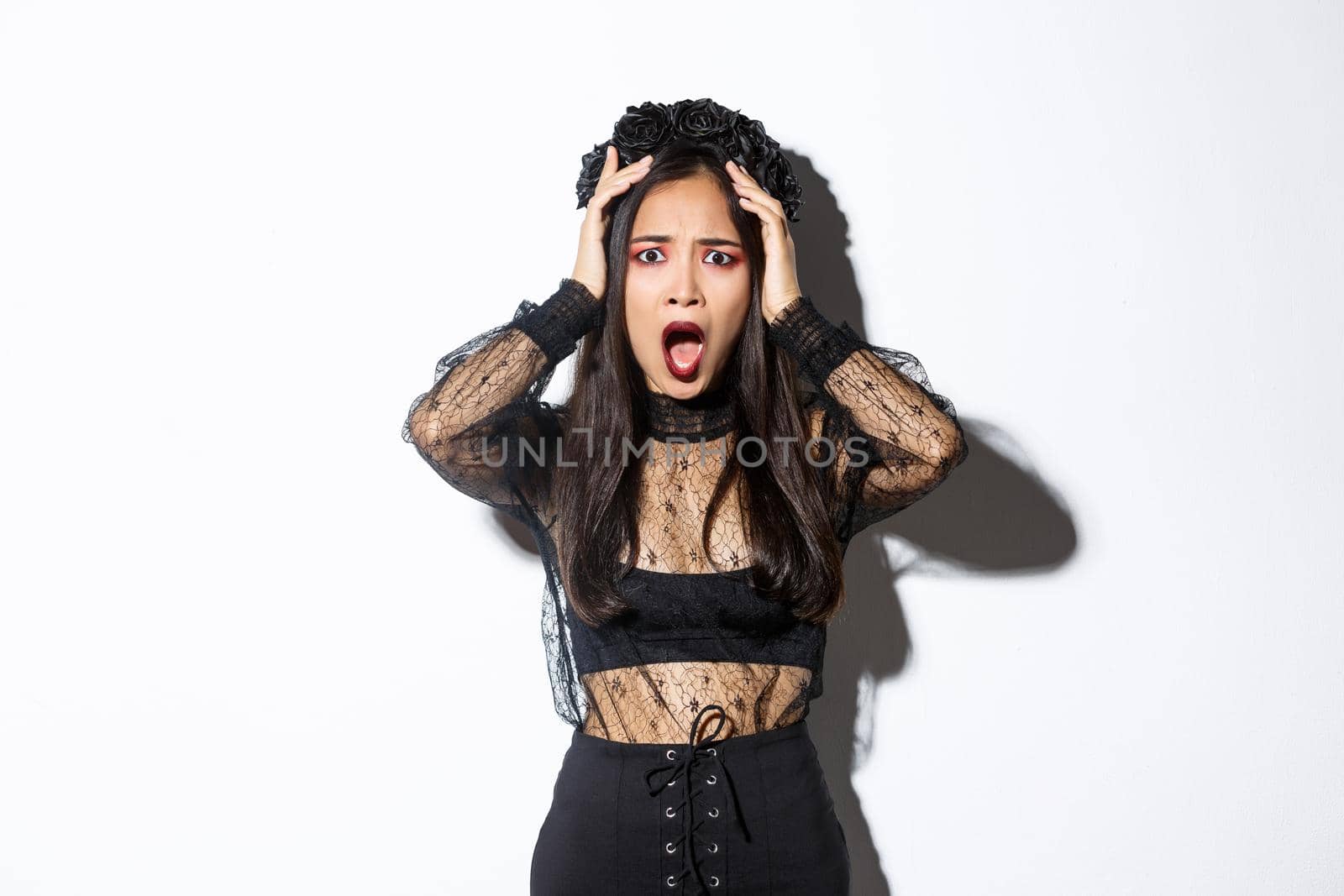 Image of horrified and shocked asian woman in gothic lace dress and wreath looking ambushed, wearing halloween costume, gasping and looking concerned, standing over white background.