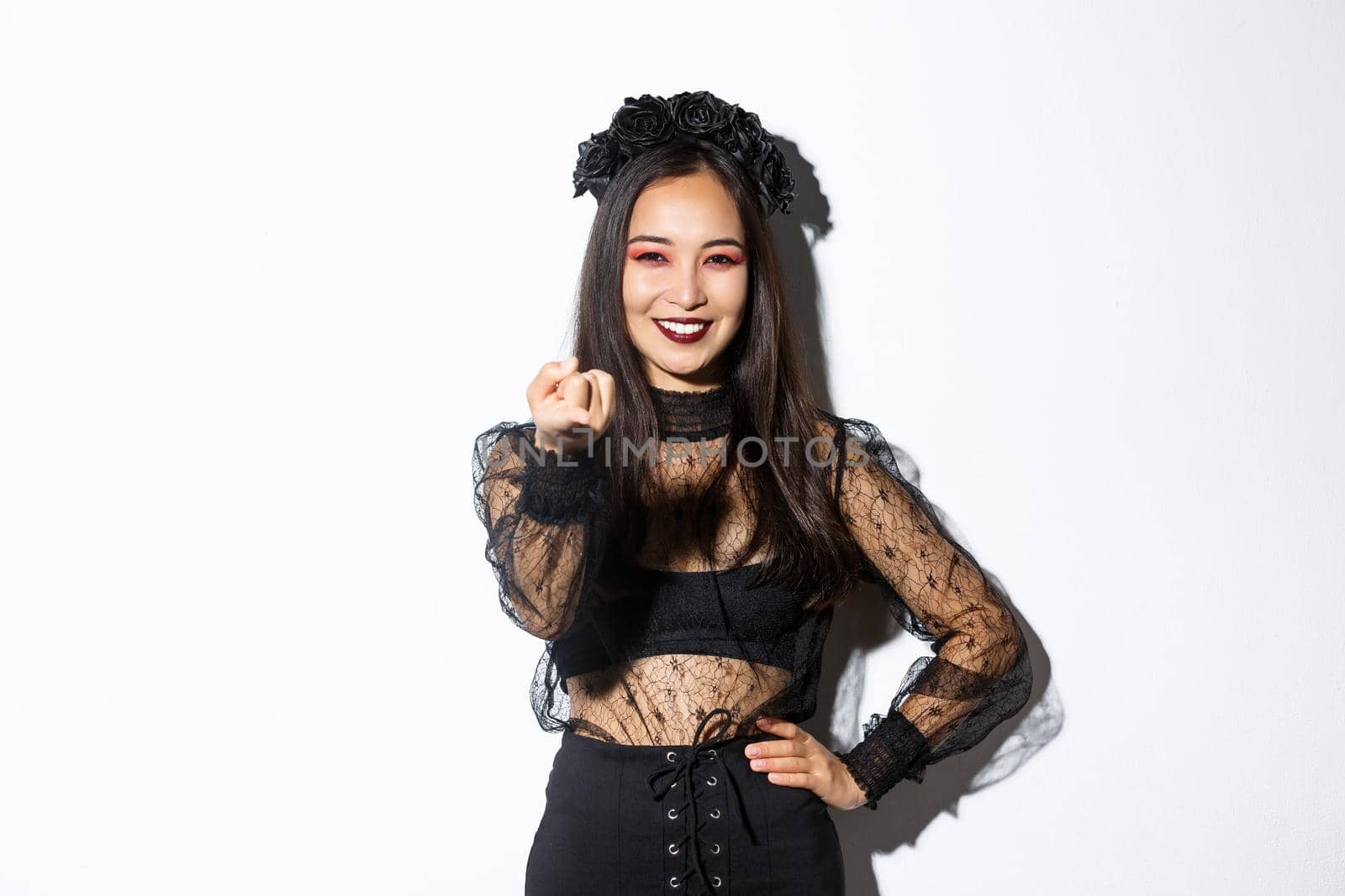 Beautiful woman in witch costume telling to come closer, flick finger and smiling, standing in halloween outfit, gothic makeup, standing over white background.