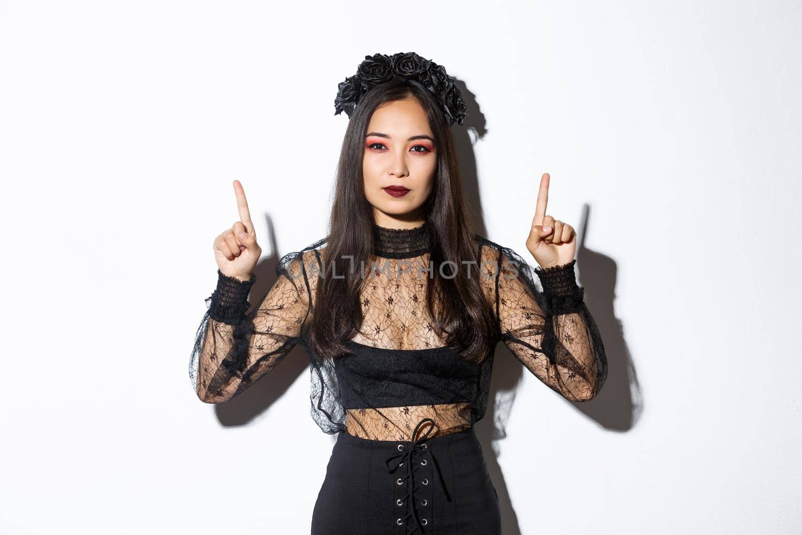 Sassy beautiful asian woman in black gothic dress, wearing witch costume for halloween and pointing fingers up, showing your logo or banner on empty white background, white background by Benzoix