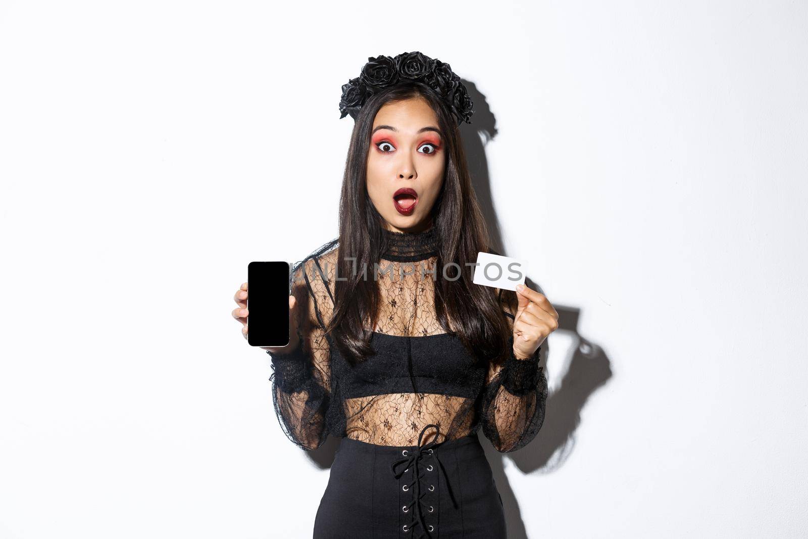 Amazed asian girl in witch costume showing credit card with mobile phone screen, open mouth surprised, celebrating halloween, white background.