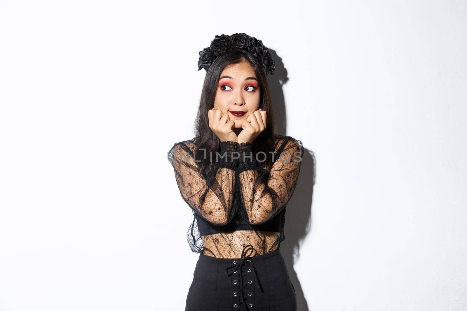Silly beautiful asian girl in gothic lace dress with wreath looking dreamy left, have nostalgic thoughts, standing in halloween costume over white background.