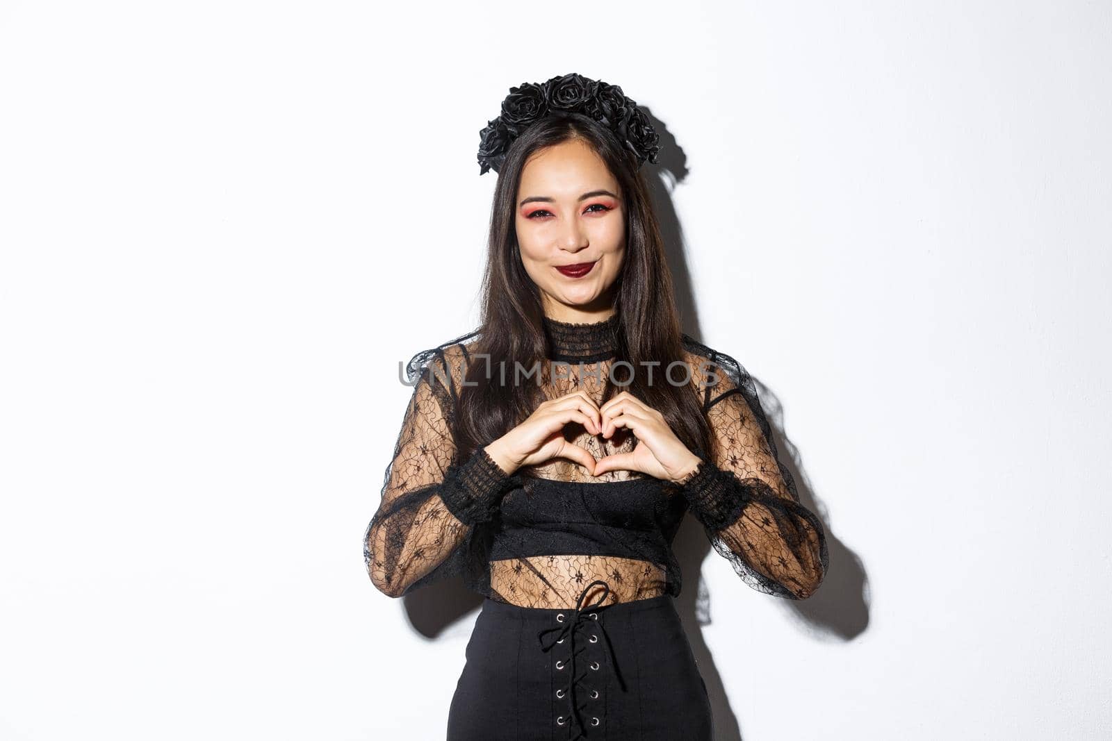 Image of lovely smiling asian woman love celebrating halloween, showing heart gesture and looking at camera, standing over white background in gothic lace dress with wreath.