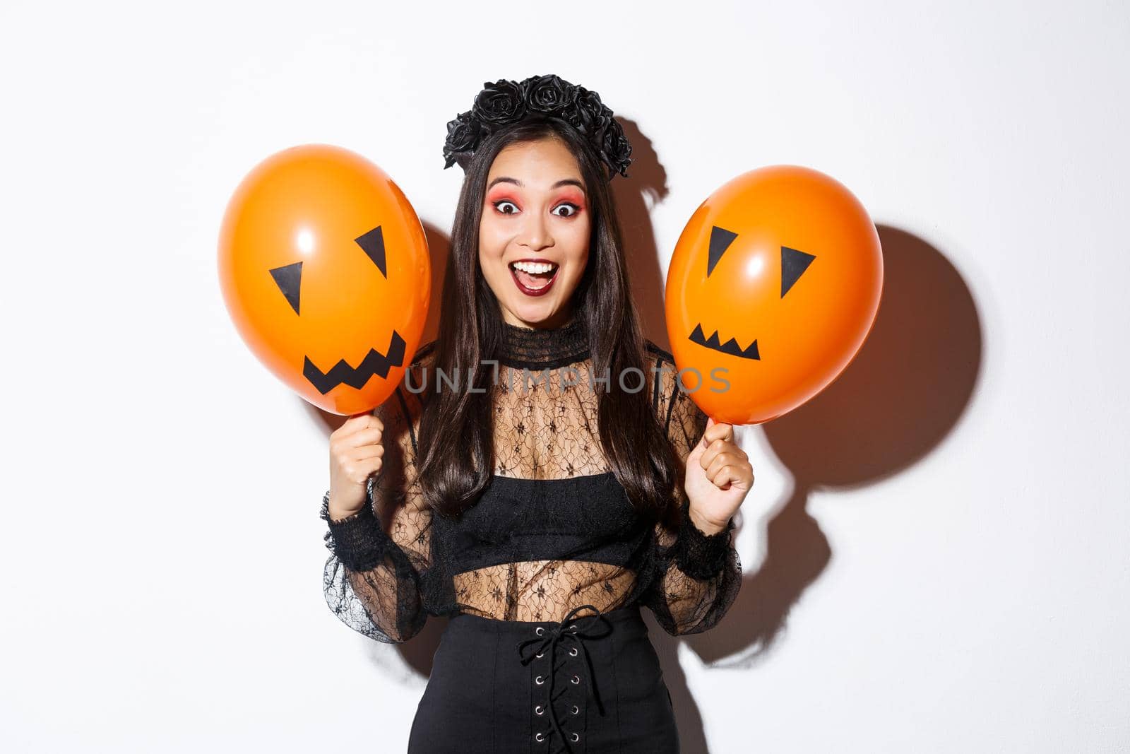 Image of happy asian woman in witch costume celebrating halloween, holding balloons with scary faces, standing over white background by Benzoix