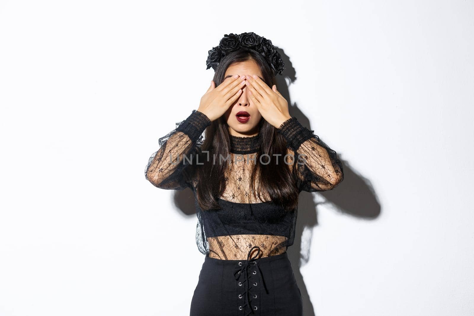 Hopeful and excited stylish asian girl in witch dress close her eyes and waiting for surprise, open mouth amused, wearing halloween costume, standing over white background.