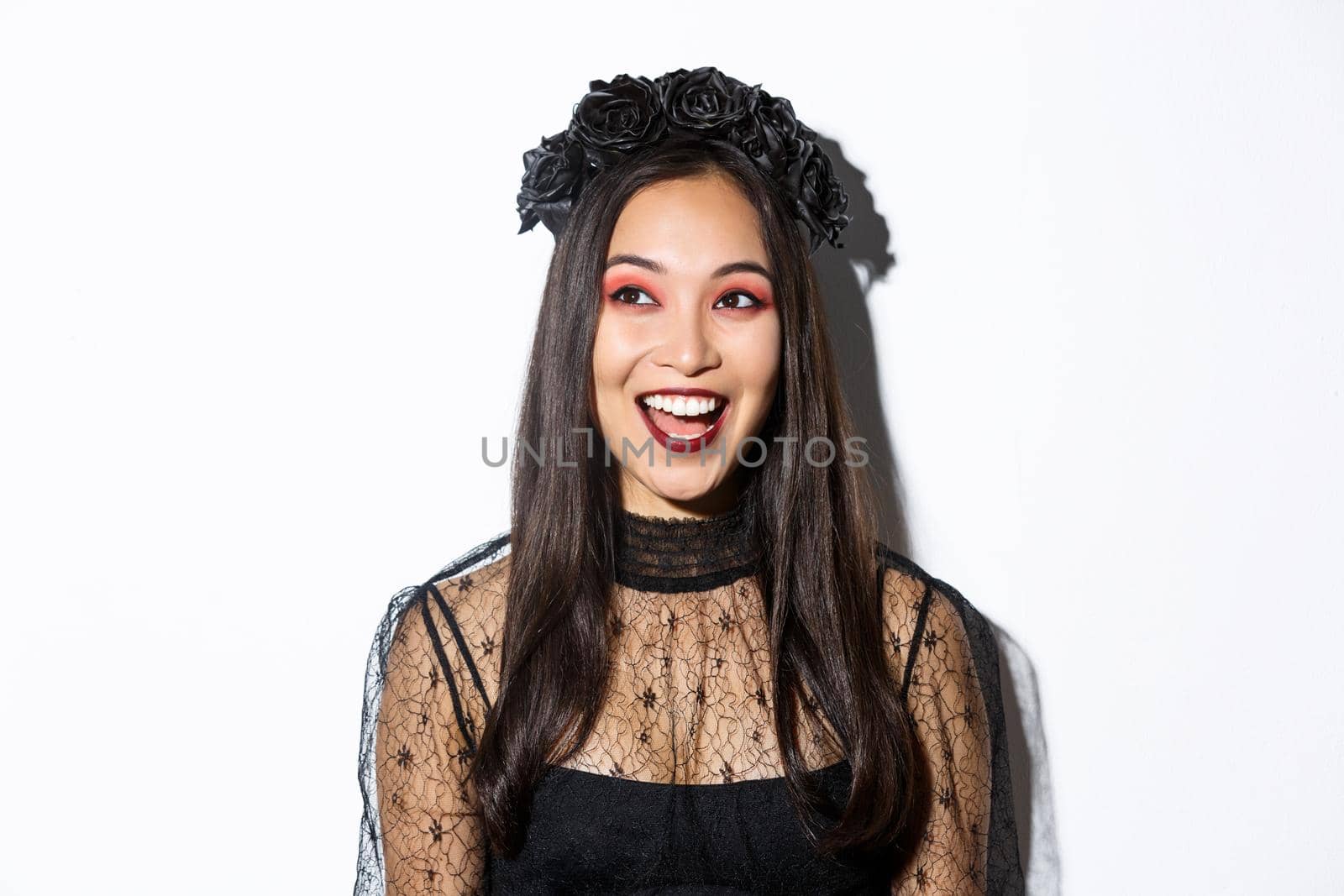 Close-up of happy pretty asian woman in halloween costume looking excited at upper left corner, standing over white background by Benzoix
