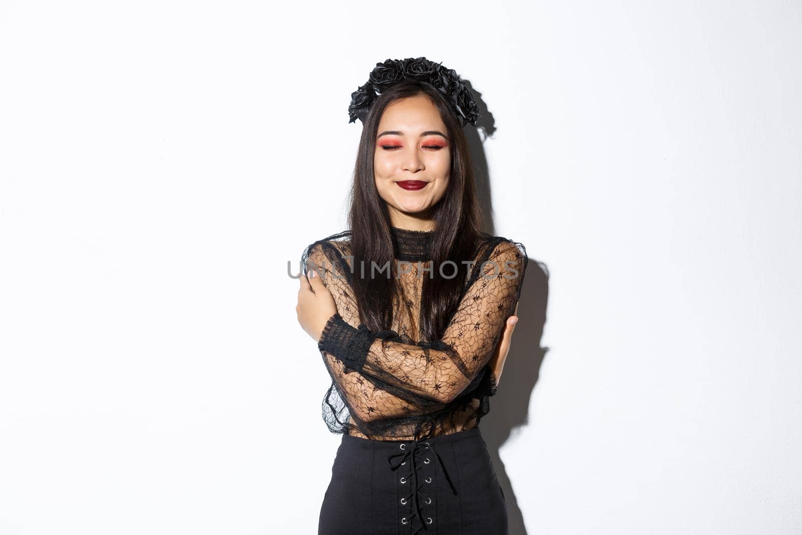 Happy cute asian woman hugging herself and smiling dreamy, wearing halloween costume of witch or widow, standing over white background.