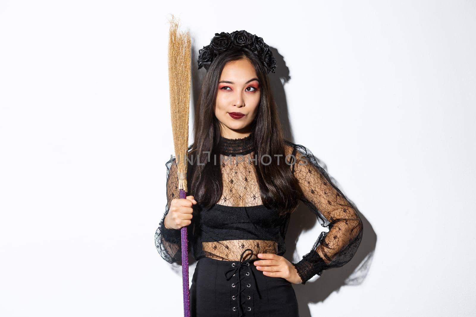 Portrait of smart thoughtful asian girl looking at upper left corner with pleased smirk, holding broom, wearing witch costume for halloween party, standing over white background.