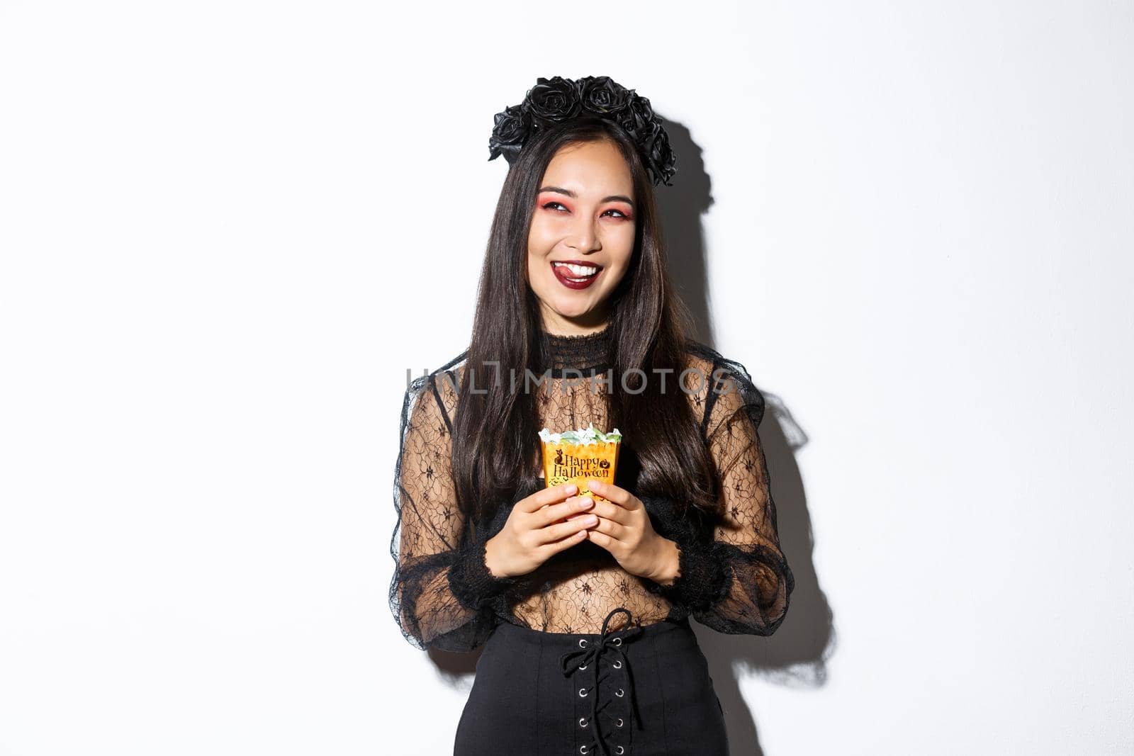 Cheerful pretty asian woman celebrating halloween in witch costume, holding sweets and smiling, looking at upper left corner cunning.