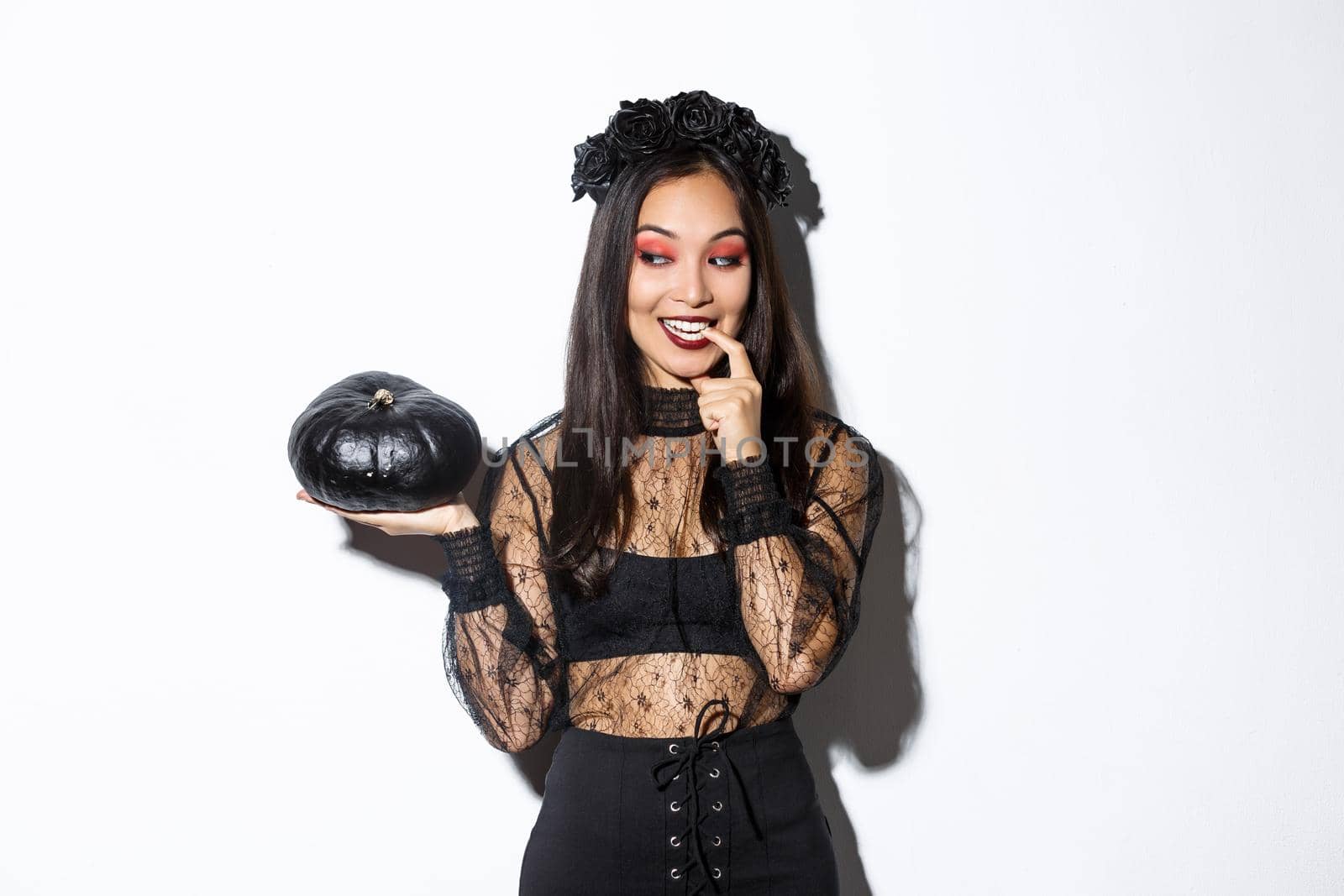 Image of sexy asian woman in witch costume looking at black pumpking and smiling, standing over white background.