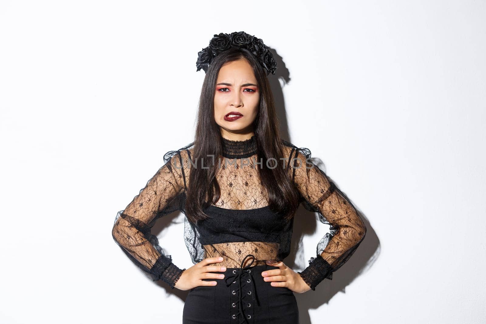 Annoyed and bothered evil witch looking complicated and biting lower lip, standing over white background in black lace dress and wreath. Girl in halloween costume trick or treating by Benzoix