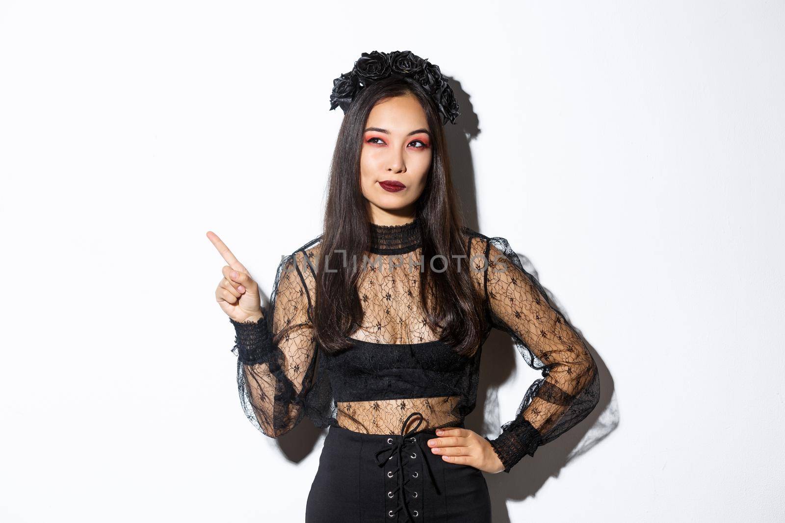 Curious attractive asian woman in witch costume, pointing finger upper left corner, looking interested in halloween promo, standing over white background and thinking.