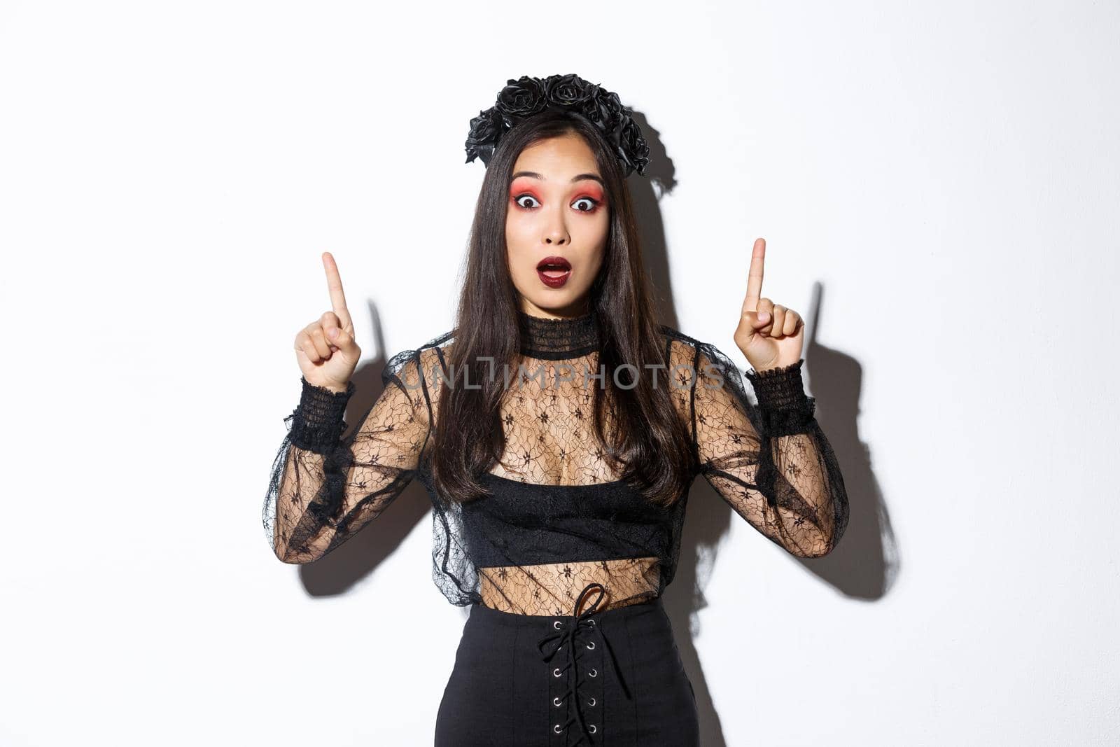 Impressed asian girl in black lace dress and wreat turn your attention on halloween promo, making announcement, pointing fingers up, showing banner, standing over white background.