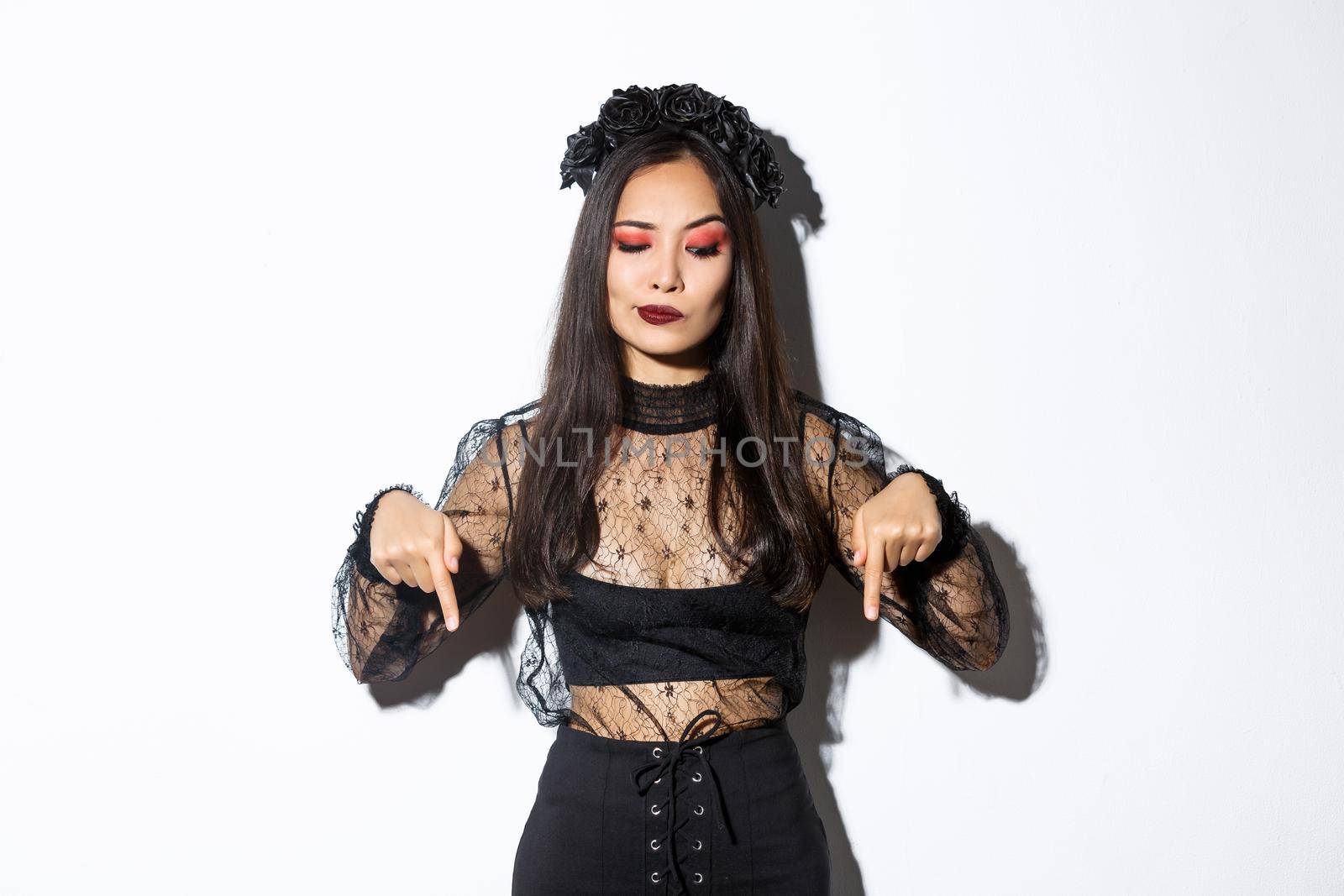 Hesitant and serious-looking asian female in magician costume, dressed-up as evil witch for halloween, looking and pointing fingers down with disbelief, standing over white background.