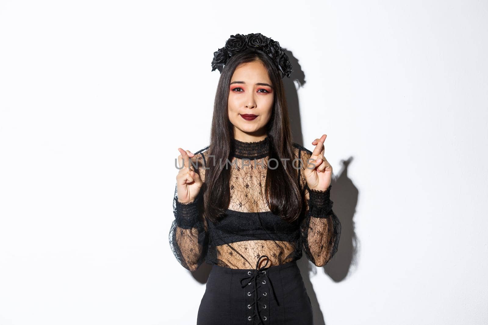 Image of hopeful asian girl in gothic lace dress and wreath making wish, cross fingers for good luck, looking wishful at camera, standing over white background by Benzoix