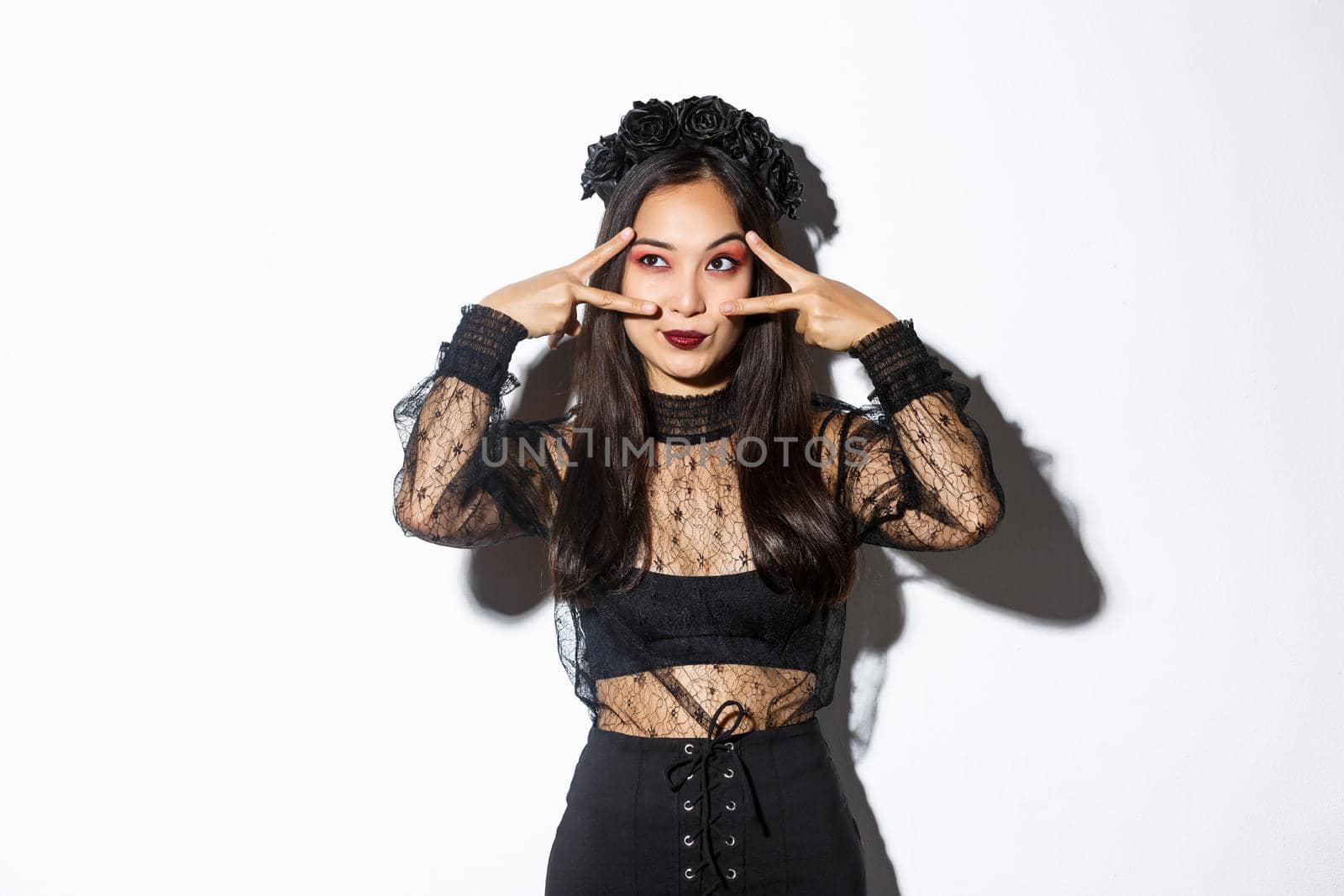 Beautiful asian woman in halloween party outfit looking at upper left corner, making peace gestures over eyes, standing in gothic lace dress with black wreath over white background.