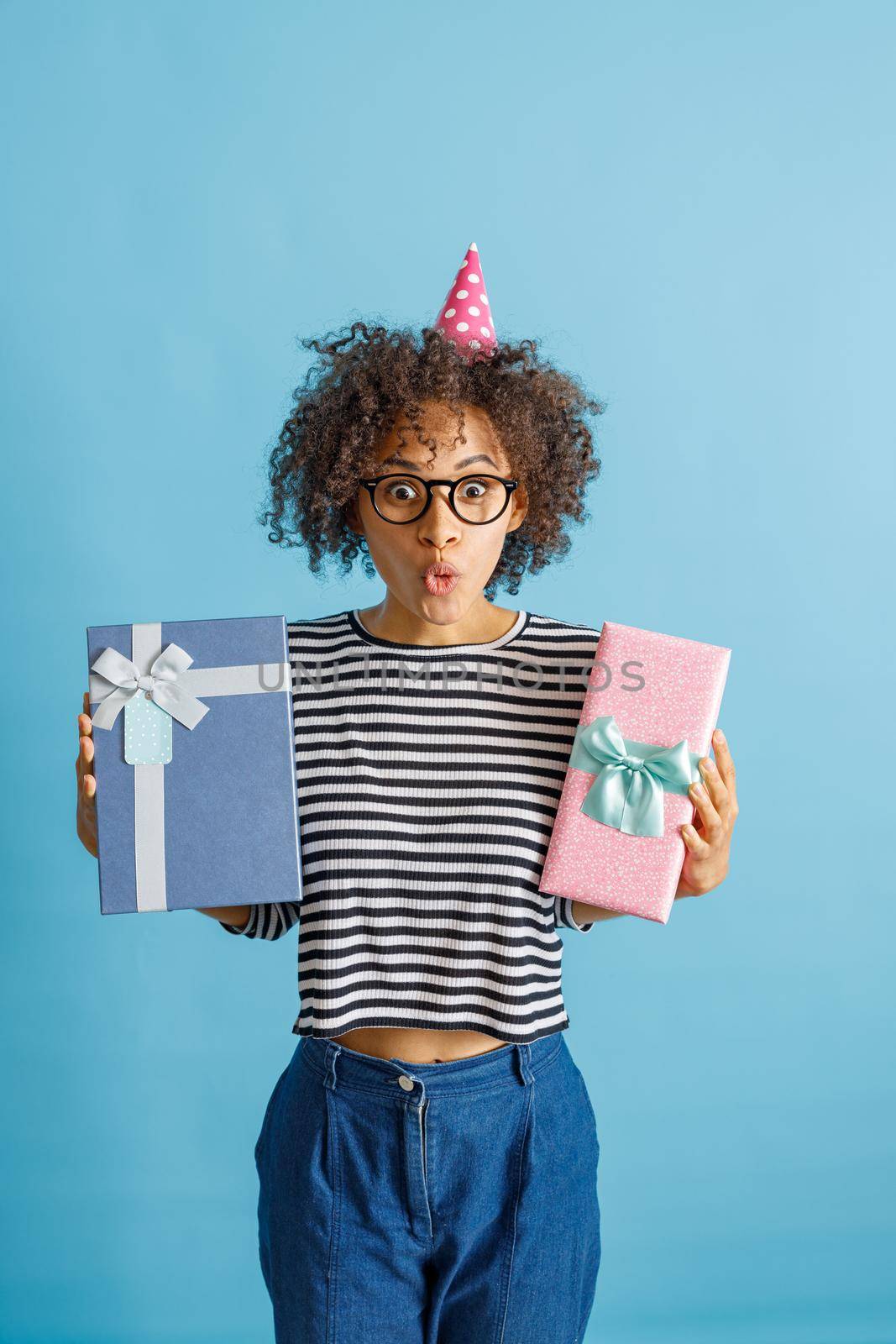 Female person in party cone hat pouting lips and making funny face while holding two wrapped gift boxes