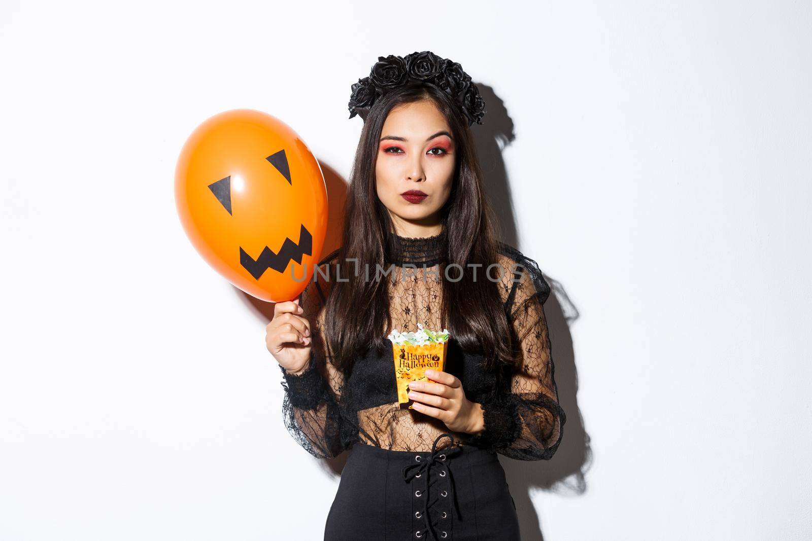 Serious-looking woman looking arrogant in witch costume, trick or treating during halloween night, holding balloon and sweets by Benzoix