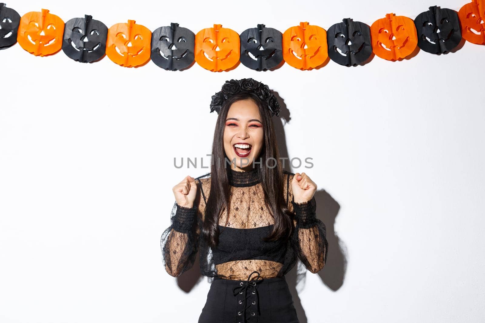 Carefree smiling asian woman in witch costume enjoying halloween party, dancing and rejoicing, standing over white background with pumpkin streamers decoration by Benzoix