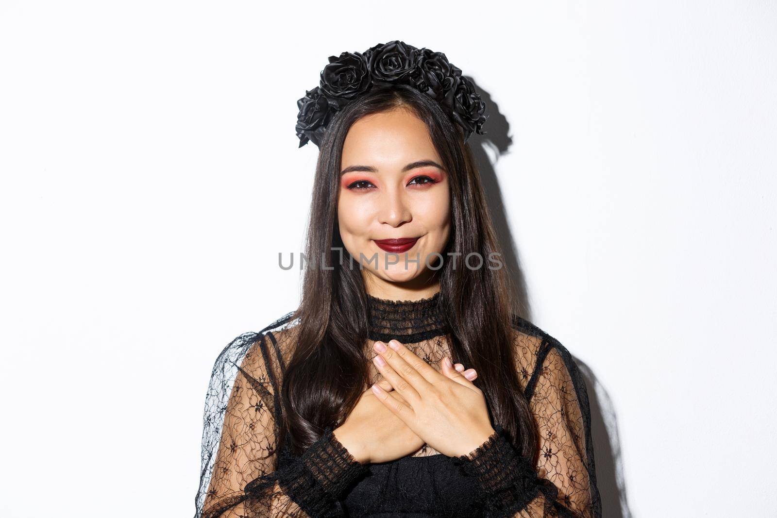 Close-up of thankful smiling asian woman looking grateful with hands over chest, standing in witch costume over white background by Benzoix