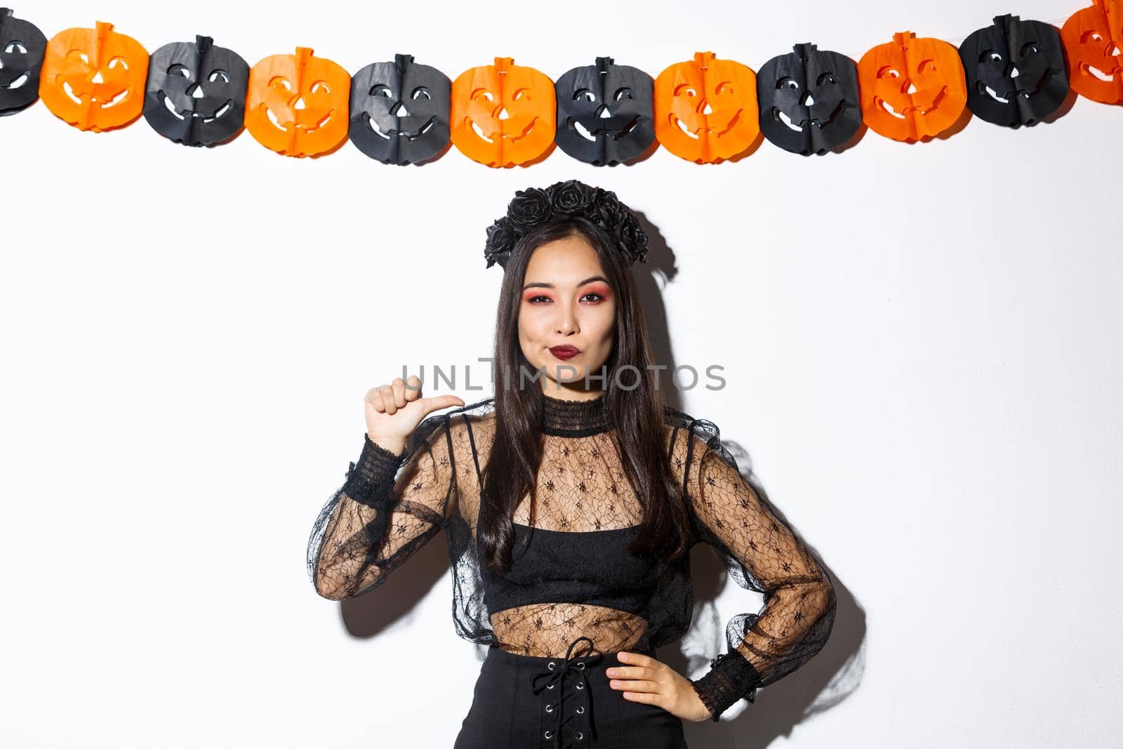 Confident asian woman in stylish gothic dress and black wreath pointing at herself like professional, standing against halloween decorations.