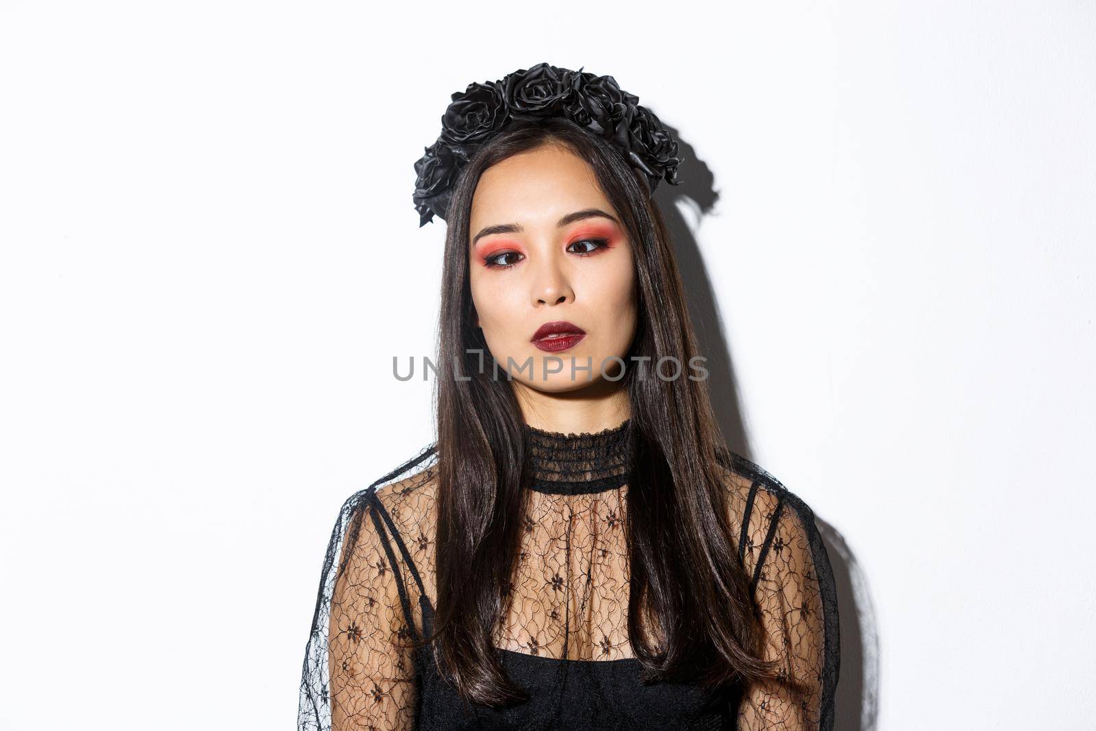 Close-up of bored and annoyed asian woman in halloween costume of witch squinting, trying to look at nose, standing over white background.