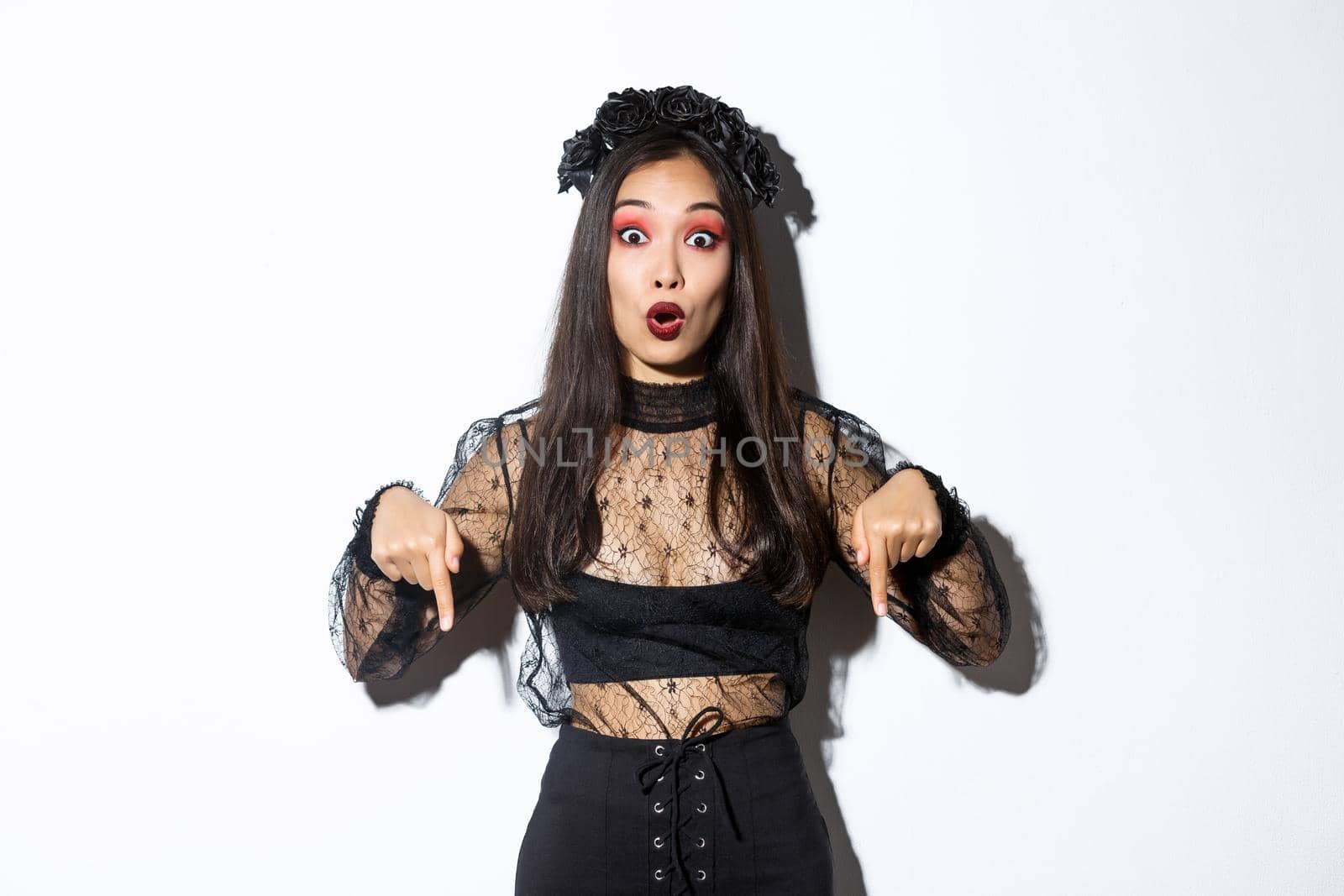 Impressed asian woman in evil witch costume pointing fingers down, showing halloween banner, making announcement. Girl wearing black lace dress and wreath, standing white background.