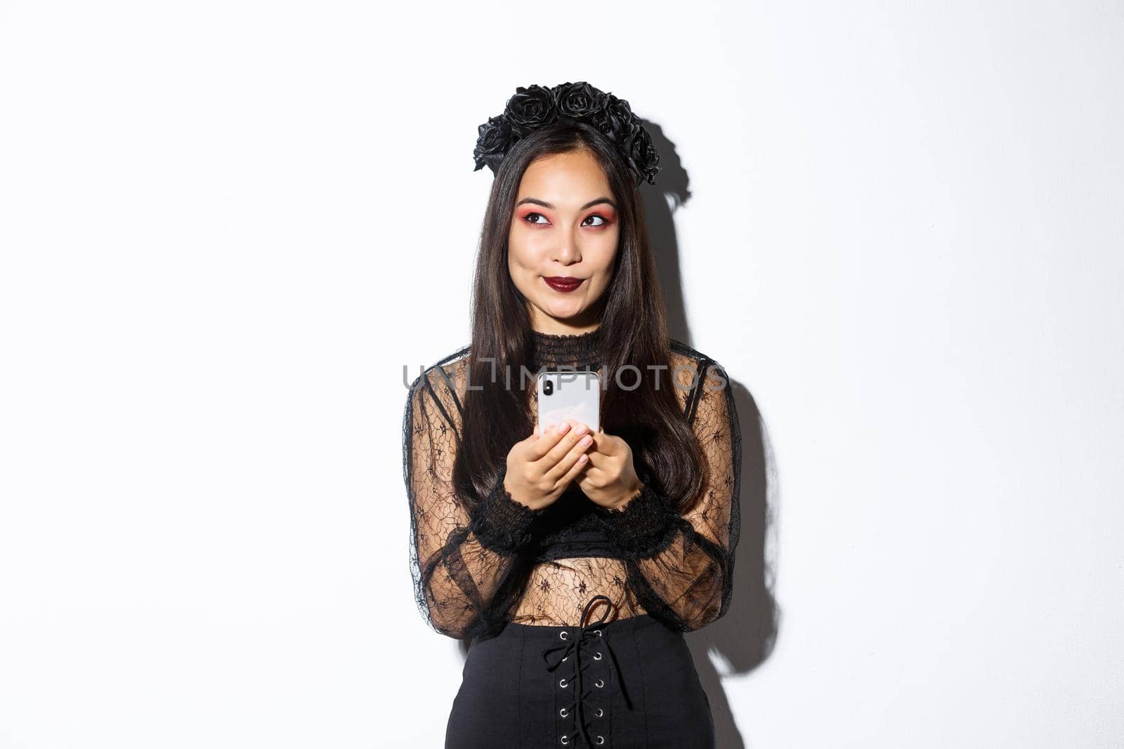 Thoughtful pretty asian girl in gothic elegant dress holding smartphone while thinking, looking at upper left corner, standing over white background by Benzoix