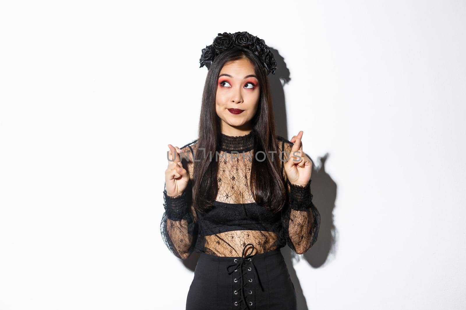 Hopeful cute asian woman in gothic lace dress and black wreath, looking upper left corner and cross fingers for good luck, making wish or praying, standing over white background.