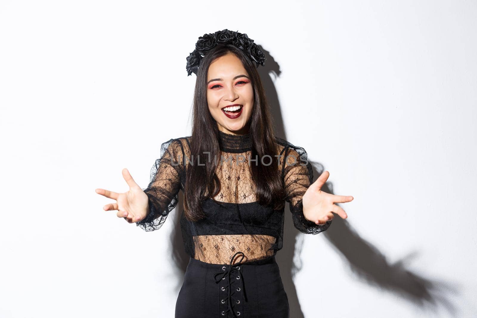 Cheerful smiling beautiful woman in black lace dress and gothic makeup inviting someone. Asian girl spread hands sideways and smiling, welcome come closer, standing white background.