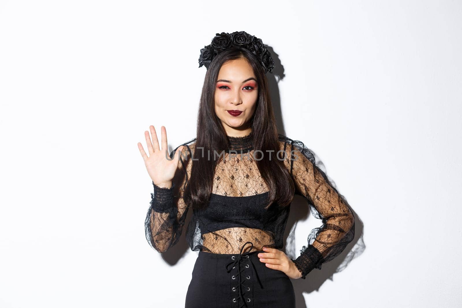 Image of confident beautiful asian woman in halloween costume showing five fingers, raising hand to say hello, standing over white background by Benzoix