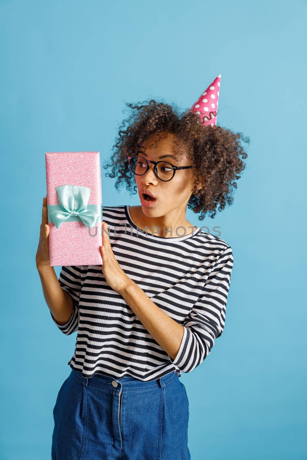 Surprised multiethnic lady in birthday cone hat holding gift box with ribbon. Isolated on blue background