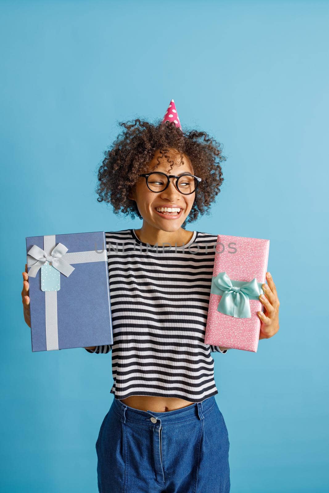 Cheerful young lady in party cone hat looking away and smiling while holding two gift boxes. Isolated on blue background